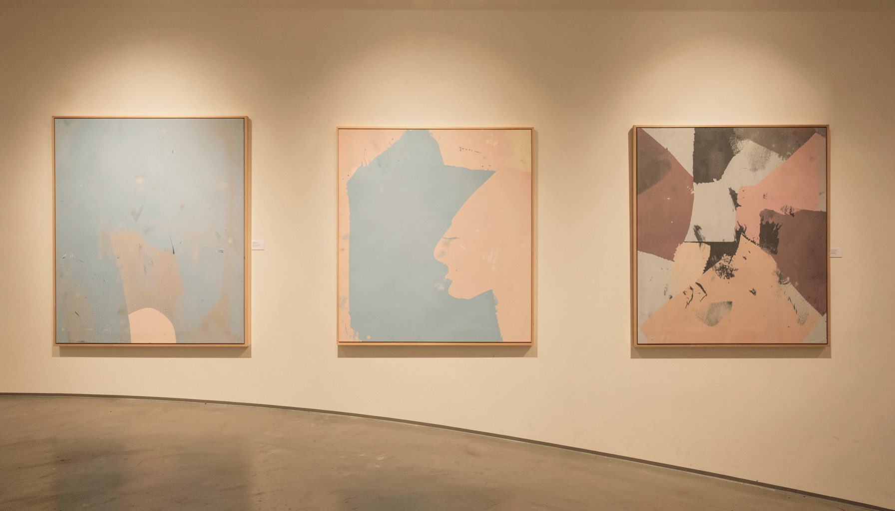 A warm, softly lit gallery wall in muted beige and cream tones, with abstract paintings in pale blue and dusty rose